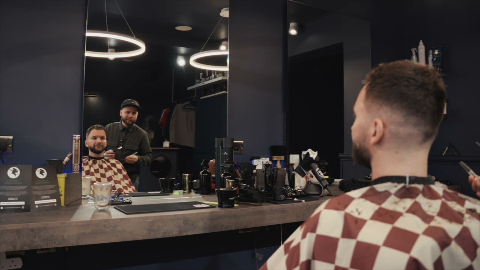 A customer in a barber's chair under ring lights, with the barber reflected in the mirror behind him